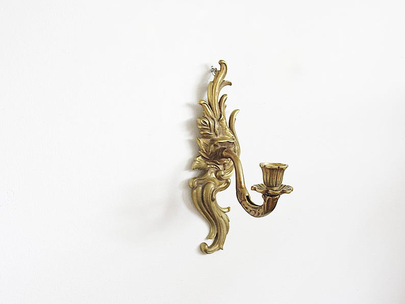 May include: Ornate brass wall sconce with a floral design. The sconce features a curved arm holding a candle cup. The decorative piece is mounted on a white wall, showcasing its intricate details and vintage aesthetic.