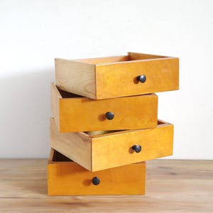 Op de afbeelding: Een stapel van vier rechthoekige houten lades met een licht oranje-bruine afwerking. Elke lade is voorzien van een enkele kleine, ronde, zwarte knop in het midden van het voorpaneel. De lades zijn in een verspringende formatie gestapeld.