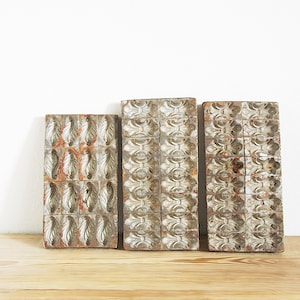 May include: Three antique, rectangular wooden molds with ornate, repeating patterns. The molds have a weathered, aged appearance with a mix of silver, brown, and orange tones. They are arranged on a light wood surface against a white background.