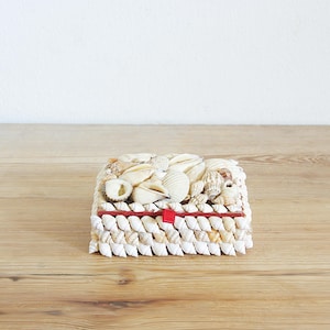 May include: A small, rectangular box made entirely of seashells. The box is filled with a variety of seashells, including white, brown, and tan shells. The box is tied with a red ribbon.