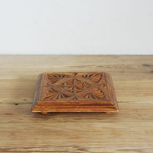 Op de afbeelding: Een vierkante houten doos met ingewikkelde geometrische patronen op het deksel. De doos heeft een warme bruine kleur en staat op kleine voetjes. De achtergrond is een lichtgekleurd houten oppervlak.