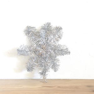 以下が含まれることがあります： 銀色の星型のクリスマスツリートッパー。トッパーは、きらきらした素材でできており、ワイヤーベースが付いています。星は、白い背景に対して、明るい茶色の木製の表面に表示されています。