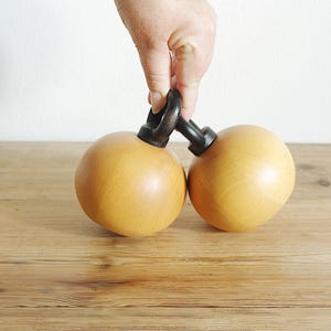 May include: Two wooden spheres connected by a black metal handle. The spheres are a light brown color and have a smooth, polished finish.