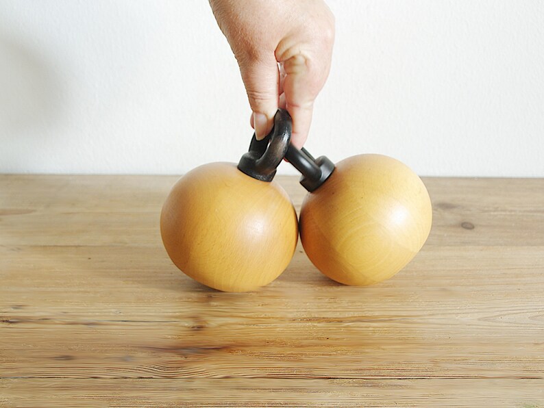 May include: Two wooden spheres connected by a black metal handle. The spheres are a light brown color and have a smooth, polished finish.