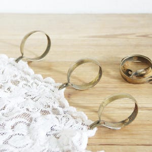 Può includere: Quattro anelli portatovaglioli antichi in metallo dal design circolare, appoggiati su una superficie di legno chiaro. È visibile una sezione di pizzo bianco, tenuta da alcuni degli anelli. Gli anelli hanno un aspetto vintage e ossidato.
