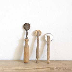 Könnte beinhalten: Drei antike Teigschneider mit Holzgriffen und Metall-Schneidrädern. Die Schneider sind auf einer Holzoberfläche vor einer weißen Wand angeordnet. Die Schneider haben unterschiedliche Raddesigns.