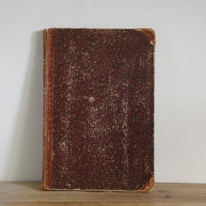 Peut inclure: Un livre ancien avec une couverture texturée et usée. La couverture est d'un brun rougeâtre foncé avec un motif tacheté. Les bords du livre montrent des signes d'usure, avec une teinte orange délavée. Le livre est posé verticalement sur une surface en bois.