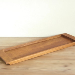 May include: A long, rectangular wooden tray with a smooth, polished finish and a warm, medium-brown color. The tray features a shallow, recessed center area with slightly raised edges on the shorter sides, showcasing a natural wood grain pattern.