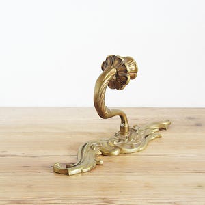 May include: Ornate brass door handle with a floral design. The handle curves upwards from a decorative base with a flowing, leaf-like pattern. The brass has a warm, aged patina, suggesting vintage or antique origins.