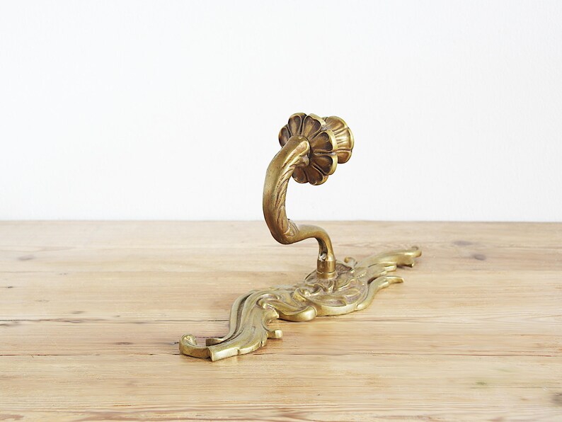 May include: Ornate brass door handle with a floral design. The handle curves upwards from a decorative base with a flowing, leaf-like pattern. The brass has a warm, aged patina, suggesting vintage or antique origins.