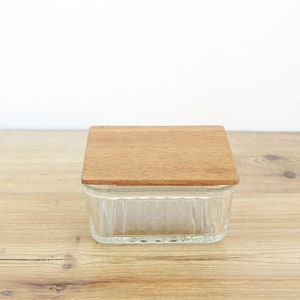 Op de afbeelding: Een glazen botervloot met een houten deksel. De vloot is rechthoekig en gemaakt van helder glas. Het deksel is van hout en heeft een natuurlijke afwerking.