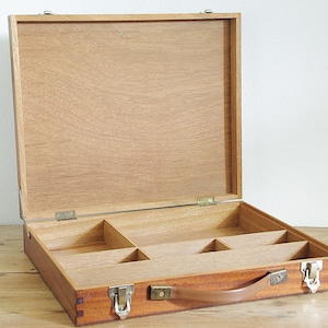 Könnte beinhalten: Geöffnete Künstlerbox aus Holz mit hellbraunem Finish. Die Box hat eine rechteckige Form mit einem Klappdeckel und Metallverschlüssen. Im Inneren befinden sich mehrere Fächer zur Organisation von Kunstbedarf. Ein brauner Ledergriff ist vorne angebracht.
