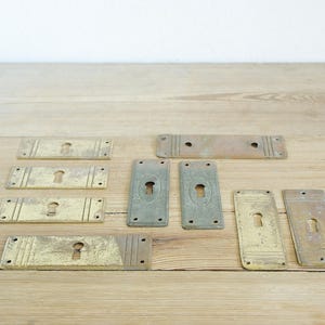 Op de afbeelding: Een verzameling van tien antieke messing sleutelgaten, sommige met een decoratieve rand, gerangschikt op een houten oppervlak.