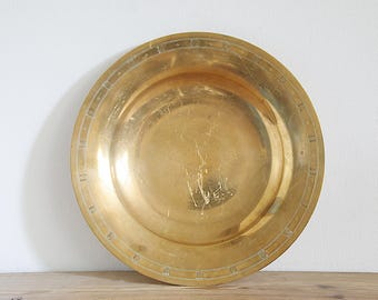 May include: A large, round, antique brass plate with a slightly concave center. The plate has a golden hue and a decorative border with small, repeating geometric shapes. The plate is displayed on a wooden surface.