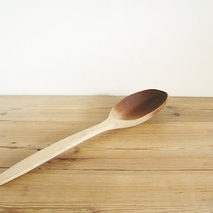 May include: A wooden spoon with a brown, worn finish. The spoon is lying on a light wood surface.
