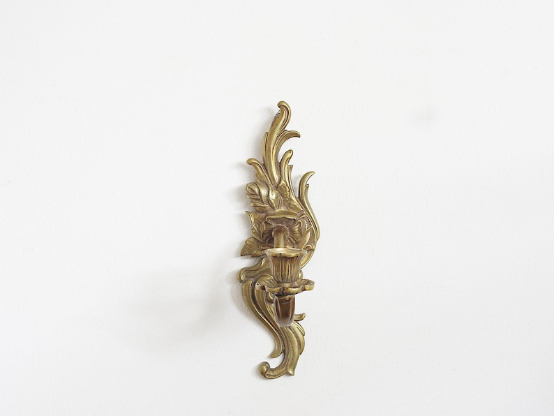 May include: Ornate brass wall sconce with a floral and leaf design. The sconce features a candle holder and decorative flourishes. The brass has a warm, aged patina. A classic piece for home decor.