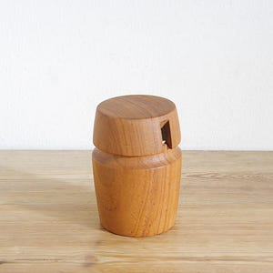 Könnte beinhalten: Ein hölzerner Salzstreuer mit zylindrischem Sockel und Deckel mit kleiner Öffnung. Der Streuer ist aus hellbraunem Holz gefertigt und steht auf einer Holzoberfläche. Der Hintergrund ist eine schlichte weiße Wand.