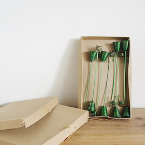 May include: A box of green metal candle holders. The candle holders are in the shape of a bell and are arranged in a row. The box is made of cardboard and is brown.