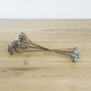 May include: A collection of vintage metal plant stakes with decorative tops. The stakes are made of thin, dark metal and have a twisted design at the top. The ends are adorned with small, gray, bead-like accents.