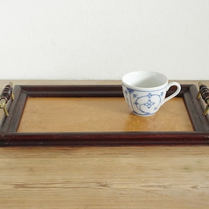 May include: A rectangular wooden serving tray with a dark brown frame and brass handles. The tray holds a white teacup with blue floral patterns. The tray is on a wooden surface.