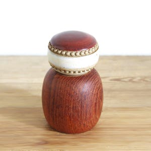Puede incluir: Un molinillo de pimienta cilíndrico de madera con un rico grano de color marrón rojizo. La sección superior presenta una tapa lisa de color marrón rojizo oscuro separada del cuerpo principal por una banda blanca con textura de cuentas en relieve.