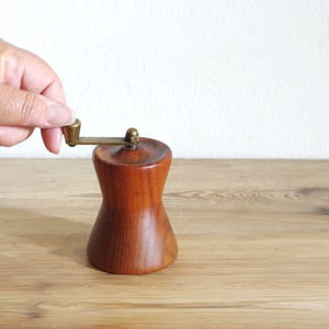May include: A wooden pepper mill with a smooth, hourglass-shaped body and a brass-colored metal crank handle on top. The wood has a warm, medium-brown tone with visible grain patterns, and the metal handle features a small, cylindrical knob.