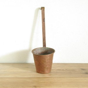 May include: A copper-colored ladle with a long handle and a small, cylindrical cup. The handle is attached to the cup with a small, rectangular piece of metal. The ladle is sitting on a wooden surface.