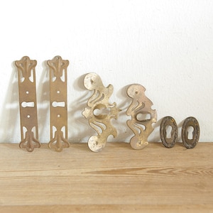 Könnte beinhalten: Eine Sammlung antiker Messingbeschläge, darunter zwei lange, verzierte Platten mit Schlüssellöchern, zwei dekorative Rosetten und zwei ovale Schlüssellochabdeckungen. Die Hardware liegt auf einer Holzoberfläche.