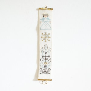 Könnte beinhalten: Ein weißer Wandbehang aus Stoff mit goldenen Verzierungen und Messingringen. Der Stoff zeigt ein Kreuzstich-Design mit einem Engel, einer Schneeflocke und einem Kandelaber.