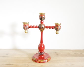 May include: A red wooden candle holder with three candle arms. The holder is decorated with gold paint and intricate floral designs.