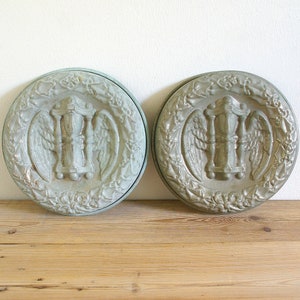 May include: Two antique, pale green, round metal molds with a raised design of an hourglass with wings. The molds are on a wooden surface.