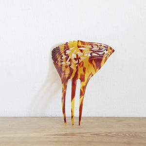 May include: A vintage hair comb with a decorative design. The comb has a tortoiseshell pattern in brown, yellow, and orange. The top section has an ornate, openwork design, and the comb has three long, pointed teeth.