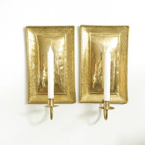May include: Two rectangular brass wall sconces with a decorative border and a candle holder. Each sconce holds a white candle. The sconces are mounted on a white wall.
