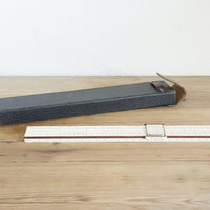 May include: A vintage slide rule with a black case. The slide rule is white with brown and black markings and a metal slider. The case is rectangular, textured, and has a leather strap. The items are on a wooden surface.