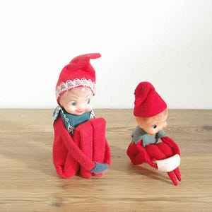 Könnte beinhalten: Zwei Elfenpuppen mit roten Filzmützen und Outfits. Die größere Puppe hat eine weiße Spitzenborte an der Mütze und einen grünen Kragen. Die kleinere Puppe hat eine schlichte rote Mütze und einen grauen Kragen. Beide haben helle Gesichter und sitzen.
