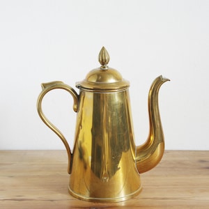 Op de afbeelding: Een vintage messing koffiepot met een gebogen handvat en tuit. De pot heeft een deksel met een kleine, decoratieve knop. Het messing is hoogglans gepolijst en reflecteert het licht. De pot staat op een houten oppervlak.