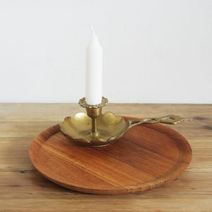 Könnte beinhalten: Ein Vintage-Kerzenhalter aus Messing mit einer weißen Kerze, auf einem runden Holztablett. Der Kerzenhalter hat ein dekoratives Blumendesign. Das Tablett ist aus Holz mit natürlicher Maserung.