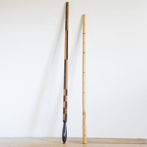May include: Two antique wooden measuring sticks. One is dark with a checkered pattern and a dark blue handle. The other is light-colored with small markings. Both are tall and slender, likely used for measuring.