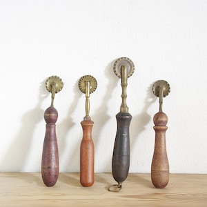 Könnte beinhalten: Vier antike Teigrädchen mit Holzgriffen und Messingklingen. Die Griffe variieren in Form und Farbe, von hell- bis dunkelbraun. Die Teigrädchen haben ein kreisförmiges, gezahntes Klingendesign.