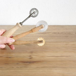 Könnte beinhalten: Drei Vintage-Teigschneider mit Holzgriffen und geriffelten Metallklingen. Die Schneider werden vor einer hellen Holzoberfläche mit weißem Hintergrund gehalten. Die Klingen haben eine kreisförmige Form mit gezacktem Rand.