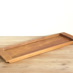 May include: A rectangular wooden serving tray with a smooth, medium-brown finish. The tray features a recessed centre area with slightly raised edges and integrated handles on both ends, showcasing a natural wood grain pattern throughout.