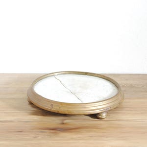 Op de afbeelding: Een ronde displaystandaard met een laag profiel, voorzien van een goudkleurige metalen rand en drie kleine pootjes. Het centrale oppervlak is een witte, ronde inleg met een opvallende, grillige barst over het grootste deel van de diameter.