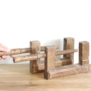 Op de afbeelding: Twee vintage houten handklemmen met dikke, verweerde bruine houten kaken en lange, schroefdraad houten spindels met cilindrische handgrepen voor handmatige aanpassing, met tekenen van ouderdom en een natuurlijke houtnerftextuur.
