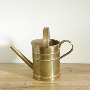 Könnte beinhalten: Eine Vintage-Gießkanne aus Messing mit langem Ausguss, gebogenem Griff und oberem Griff. Die Kanne hat einen zylindrischen Körper mit horizontalen Bändern und einem Deckel. Sie steht auf einer Holzoberfläche.