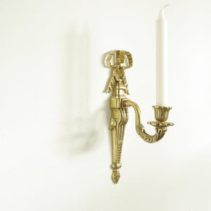 Può includere: Un portacandele da parete dorato con dettagli decorativi e una singola candela bianca.