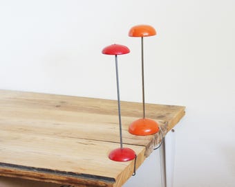 May include: Two hat stands, one red and one orange. Each has a circular base and a domed top, connected by a thin, dark metal rod. They are displayed on a wooden table. The table is made of wood.