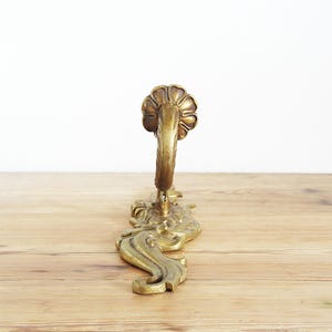 May include: Ornate brass wall hook with a floral design at the top and decorative scrollwork. The hook is a warm gold color and is mounted on a wooden surface. The hook is designed for hanging items.