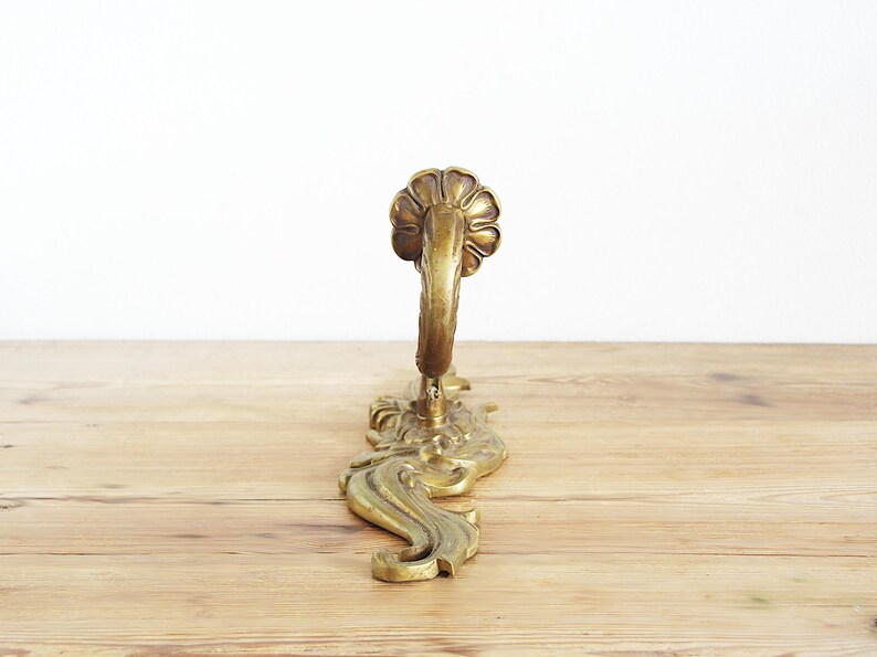 May include: Ornate brass wall hook with a floral design at the top and decorative scrollwork. The hook is a warm gold color and is mounted on a wooden surface. The hook is designed for hanging items.