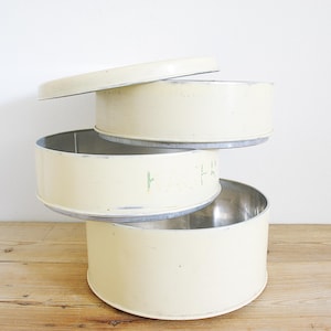 Op de afbeelding: Drie vintage, crèmekleurige cakeblikken gestapeld op een houten oppervlak. De blikken hebben een cilindrische vorm met een deksel erop. De binnenkant van de blikken is zilver. Het woord "FR" is zichtbaar op één blik.