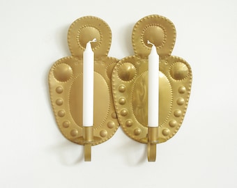 May include: Two gold-colored metal wall sconces, each holding a white candle. The sconces have a decorative, textured design with a series of small, raised circles. The candles are white and cylindrical.